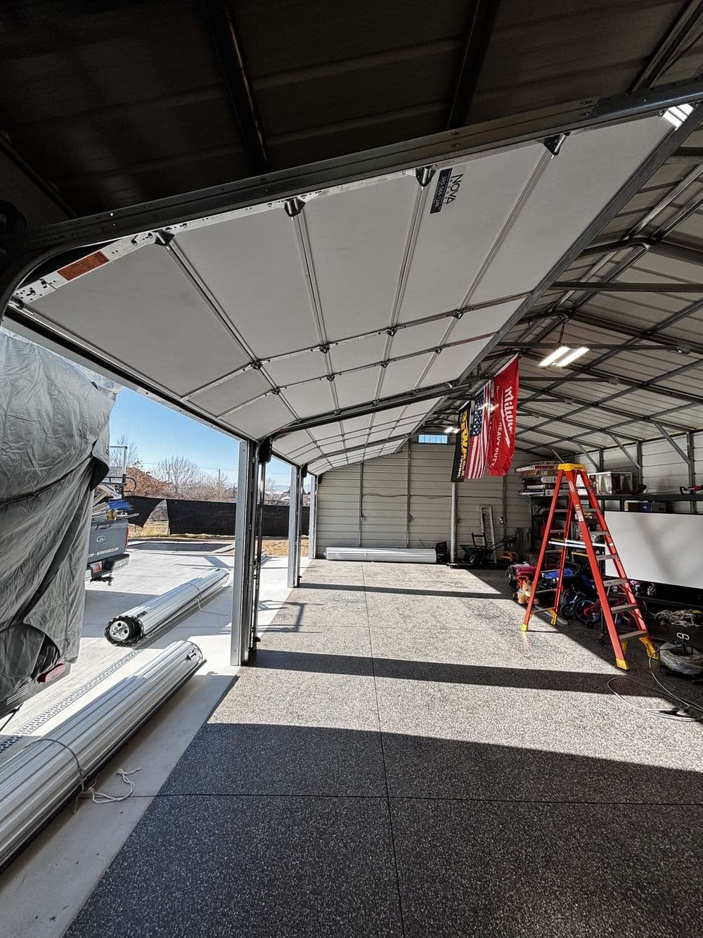 Spacious garage interior with open doors, ladder, flags, and organized storage area.