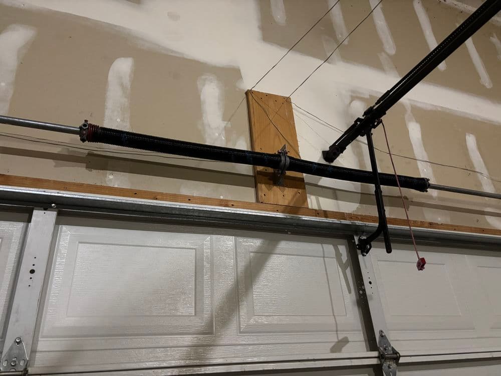 Garage door spring mechanism with black coil spring mounted on wooden board.