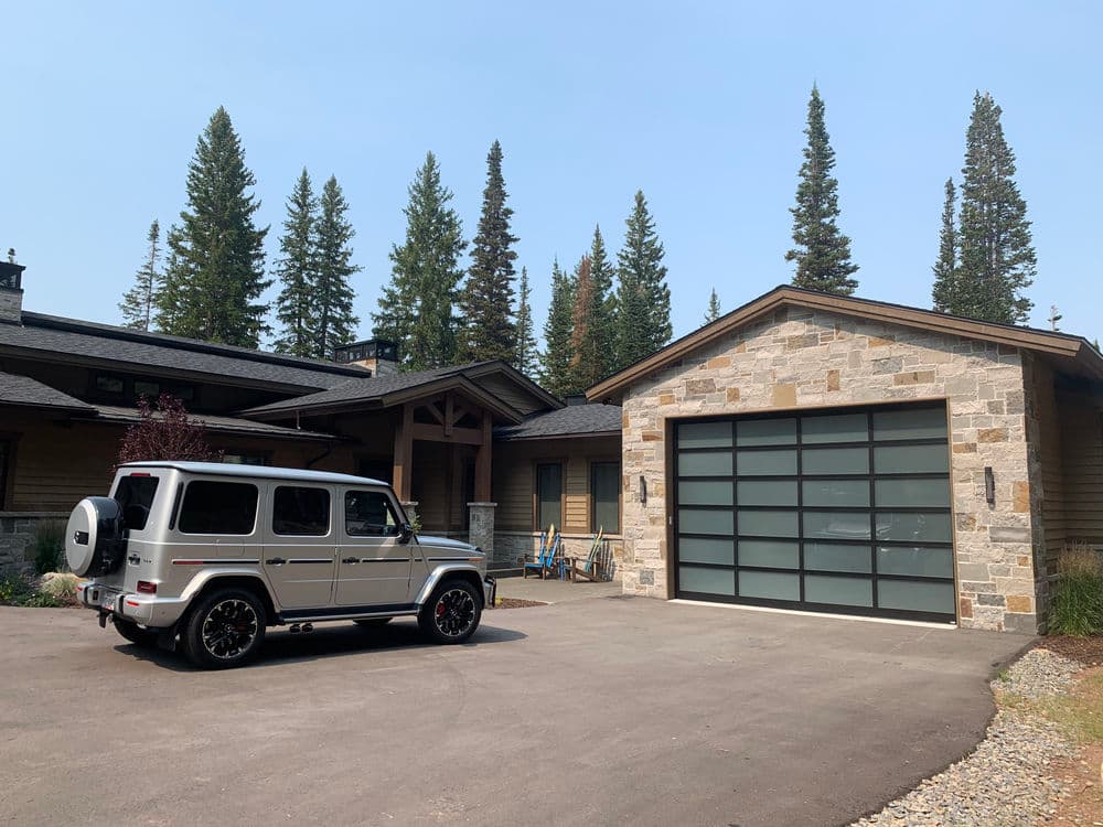 Silver SUV parked in front of modern home with stone garage, surrounded by tall evergreen trees.
