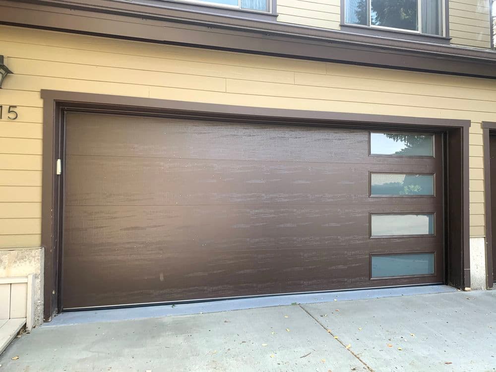 Modern brown garage door with glass panels and textured finish, set against a tan house exterior.