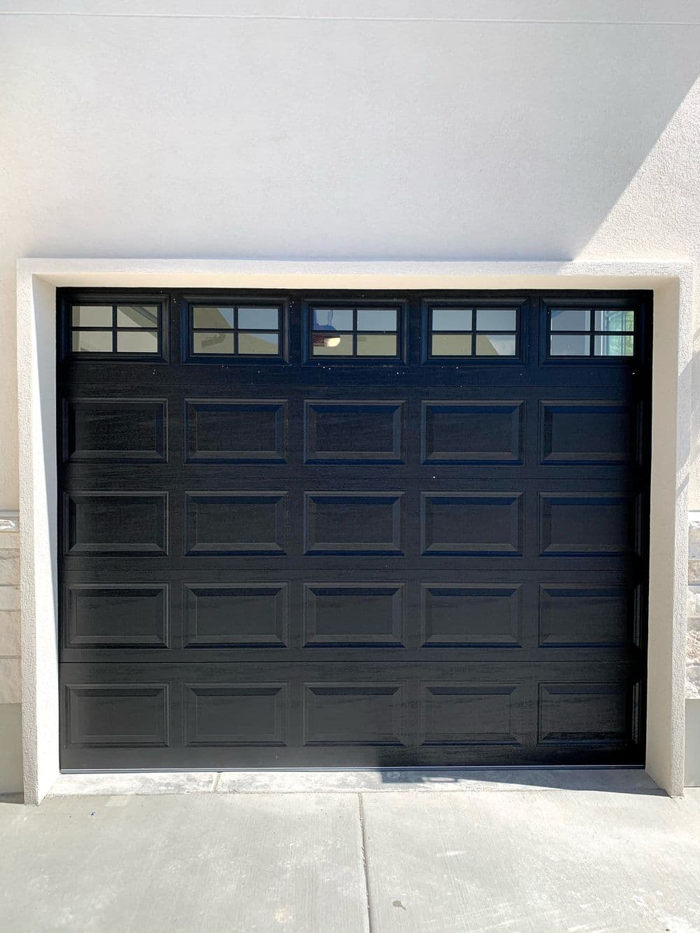 Black garage door with rectangular panels and windows, set against a light-colored wall.