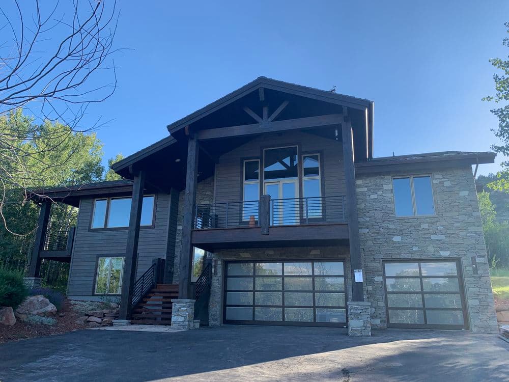 Modern stone and wood house with large windows and a three-car garage surrounded by trees.