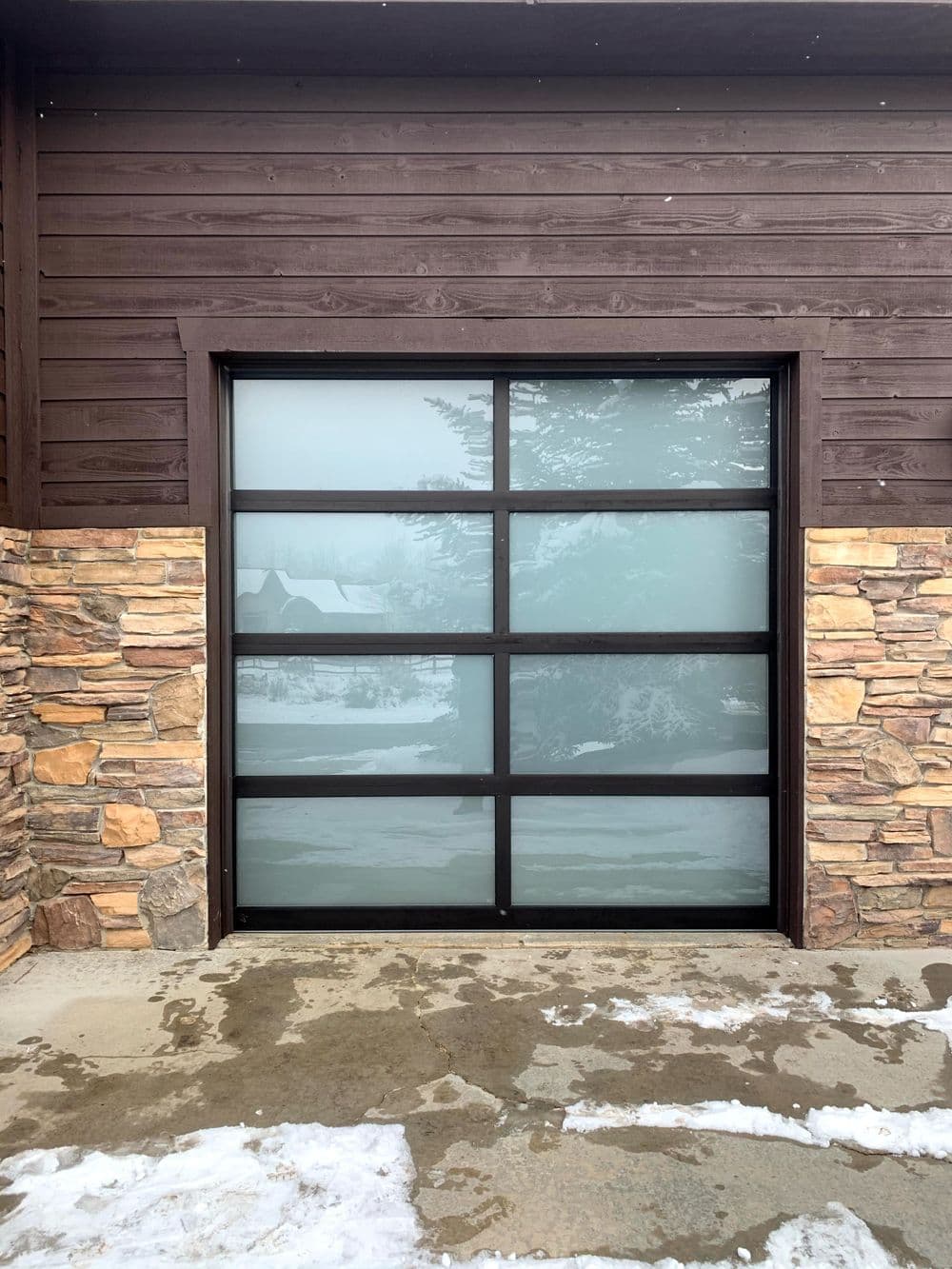 Modern glass garage door with frosted panels and stone accents on wooden exterior.