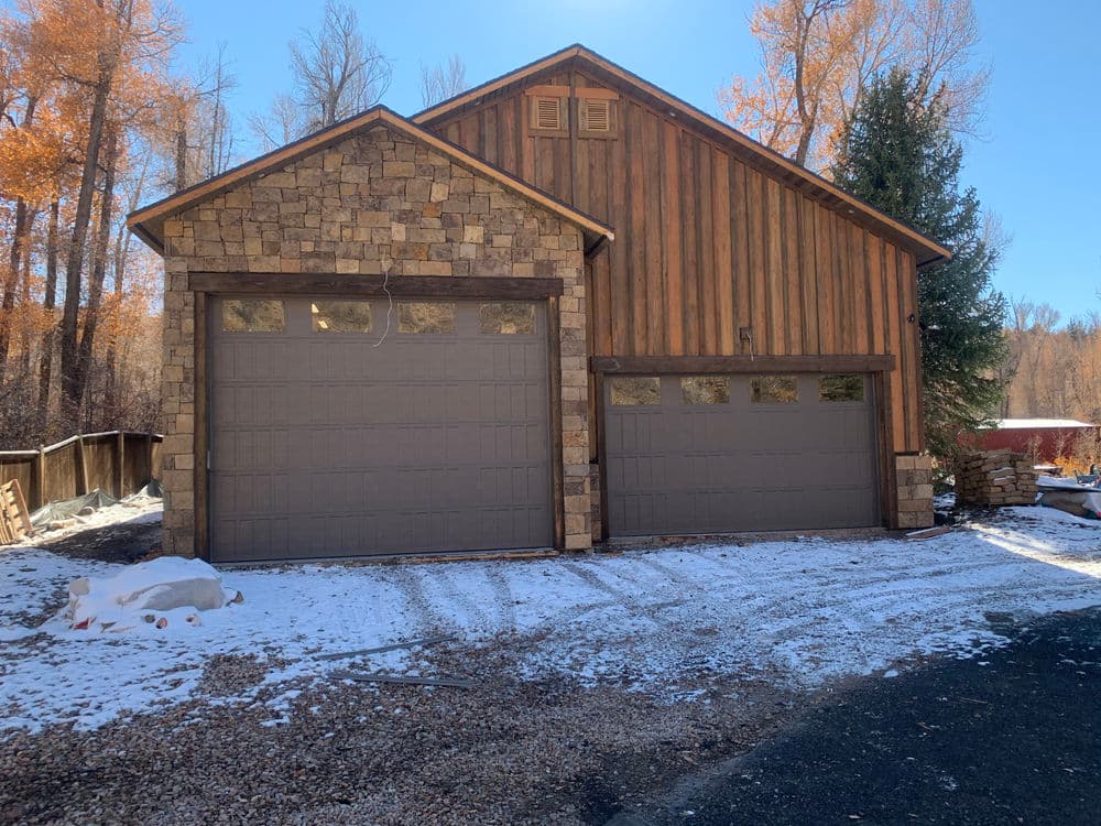 Rustic wooden house with stone accents and two garage doors in a snowy landscape.