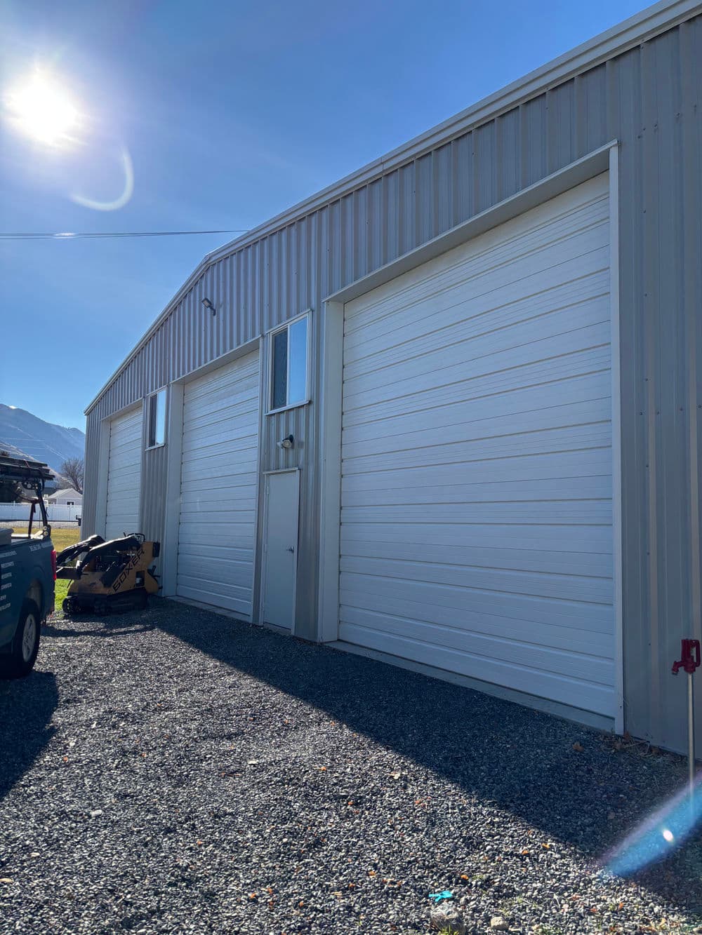 Metal storage building with large roll-up doors and gravel driveway under bright sunlight.