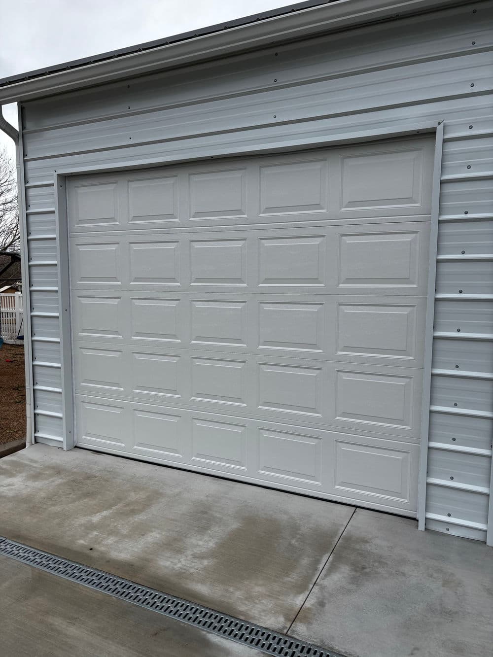 White garage door on a metal building with a concrete driveway and drainage channel.