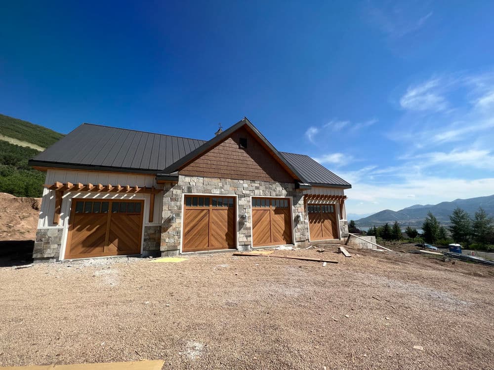 Newly constructed mountain home exterior with stone and wooden garage doors under a clear blue sky.