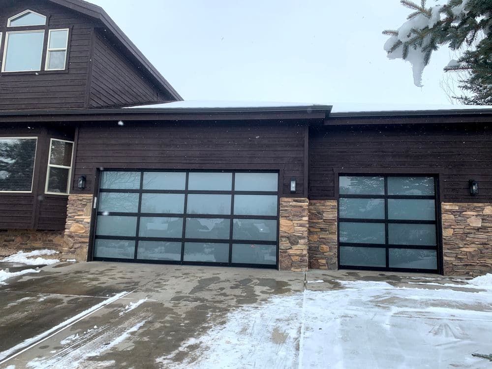 Modern two-car garage with large glass panels, stone accents, and snow-covered driveway.