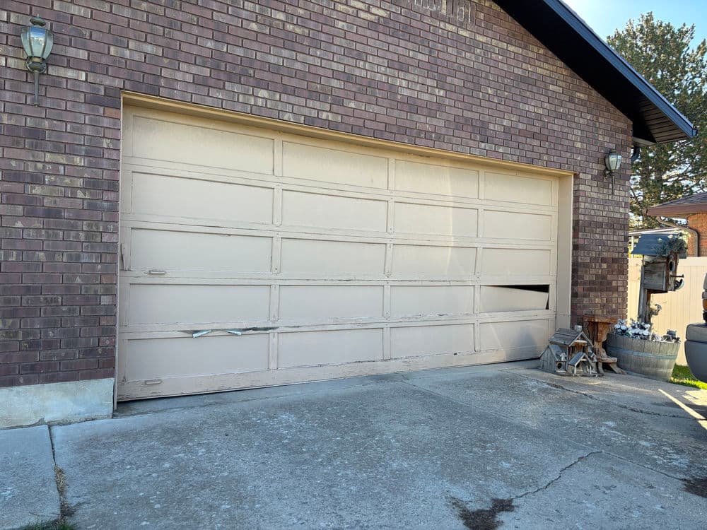 Damaged beige garage door on brick house with concrete driveway and decorative planters.