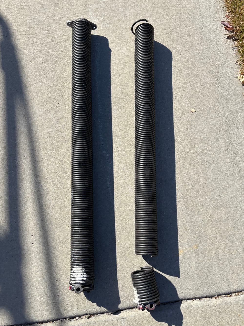 Two black garage door springs on a concrete surface, one is intact and the other is broken.