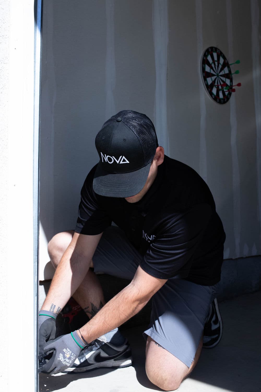Technician wearing NOVA cap installs equipment in a garage with dartboard background.