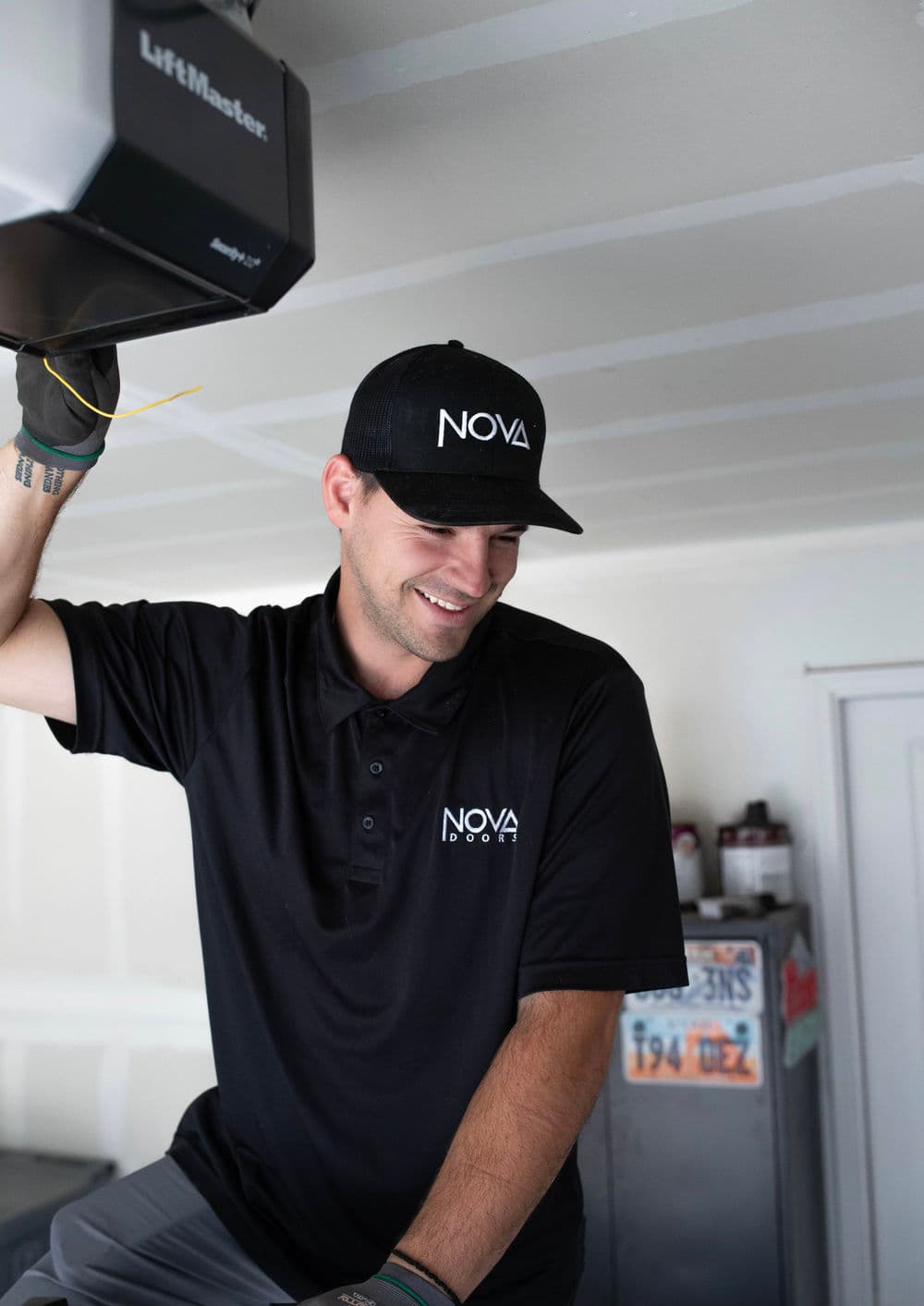 Smiling technician in a black polo and cap with "NOVA" logo working in a garage.